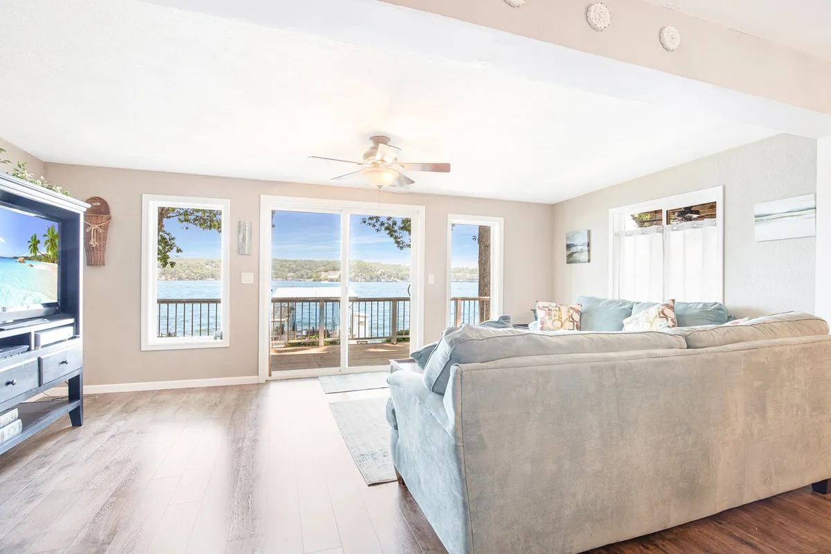 A spacious living room featuring large windows that provide sweeping views of the lake. The comfortable seating area faces both the TV and the water, allowing for relaxation and entertainment amidst abundant natural light. The decor is tranquil, with soft tones and modern furnishings.