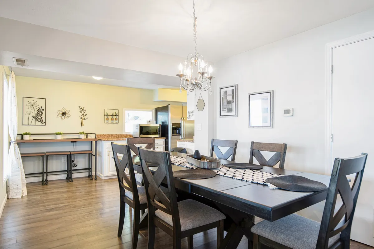 A warm, inviting dining area with a family-sized dining table under a chandelier, wall art, and an open view toward the kitchen bar. The arrangement encourages group meals and social gatherings.