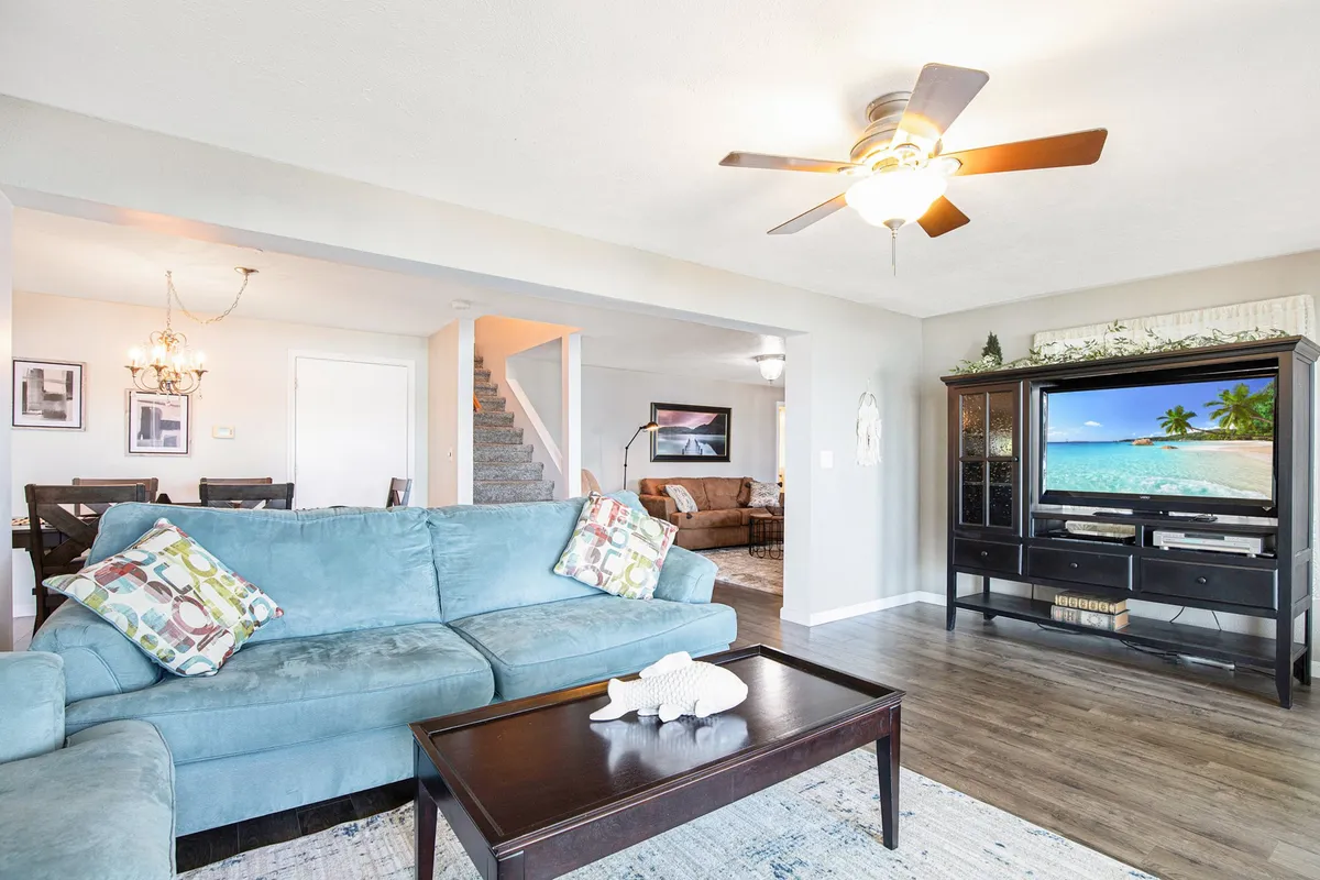 A bright, open-plan living room with a cozy sofa, modern coffee table, TV, dining area, and staircase. The space is welcoming and ideal for both relaxation and socializing, with ample natural light and well-coordinated decor.