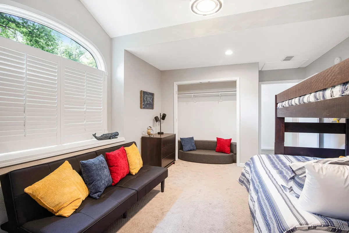 Inside, a playful bunk bed shares space with a cozy sofa adorned with colorful cushions, and a large window brightens the room. The inviting setting is tailored for children or family sleepovers, supporting both fun and relaxation for all ages.
