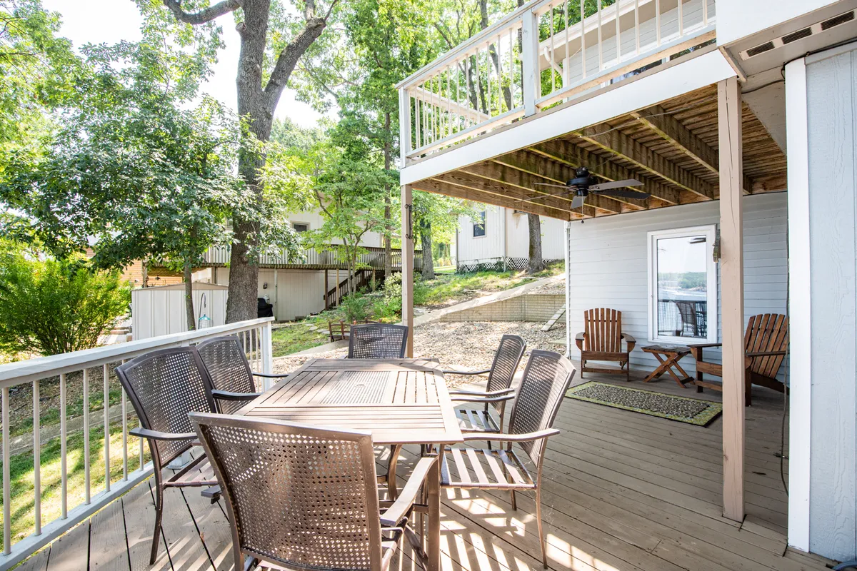An exterior shot of a comfortable shaded deck furnished with a wooden dining table, plush seating, and surrounded by lush greenery. The scene is set for outdoor dining or relaxation, with a tranquil, casual vibe that invites guests and their pets to unwind after a day of lakeside exploration.